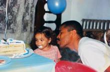Photo of a father blowing out a candle on a birthday cake. Beside him is his young daughter, smiling, her mouth covered in frosting. 