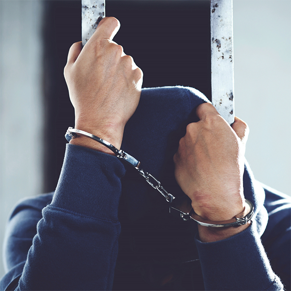A man with a sweatshirt hood pulled over his head obscuring his face stands behind bars, his hands in handcuffs gripping the bars.