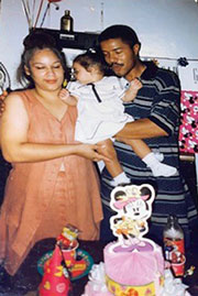 A photo of Leonel, his wife, and daughter, at a birthday celebration, with balloons behind them and a cake and gifts on a table in front of them